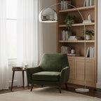 Beside a wooden bookshelf, the Clérachic arc floor lamp's chrome dome shade reaches over a green velvet armchair.