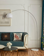 Its marble base visible on the wood floor, the Clérachic arc floor lamp arches behind the teal sofa with a white dome shade.