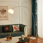 Clérachic stainless steel arc floor lamp curving over a teal tufted sofa, with whiskey glasses set on round marble coffee tables.