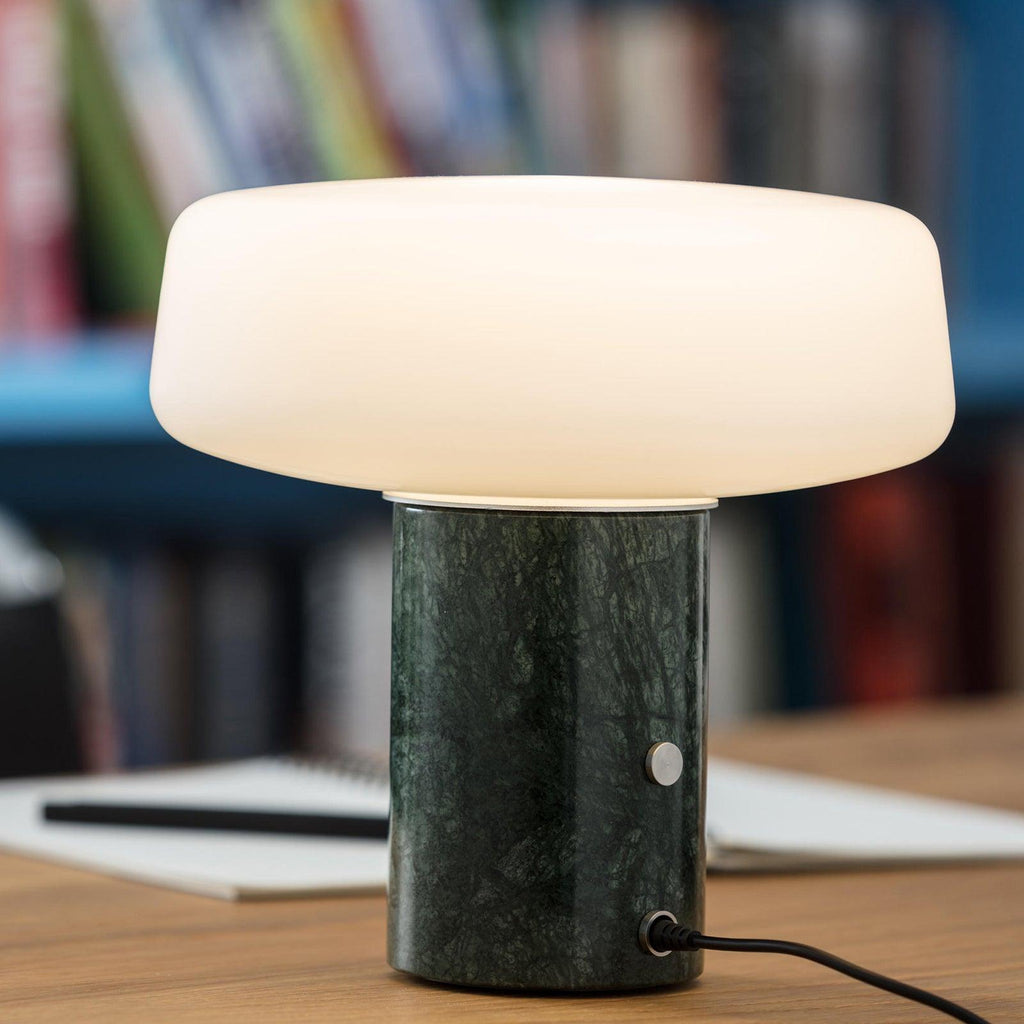 Green marble Clérachic table lamp lit on a wooden desk, its power cord and round switch visible against a blurred bookshelf.