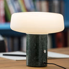 Green marble Clérachic table lamp lit on a wooden desk, its power cord and round switch visible against a blurred bookshelf.