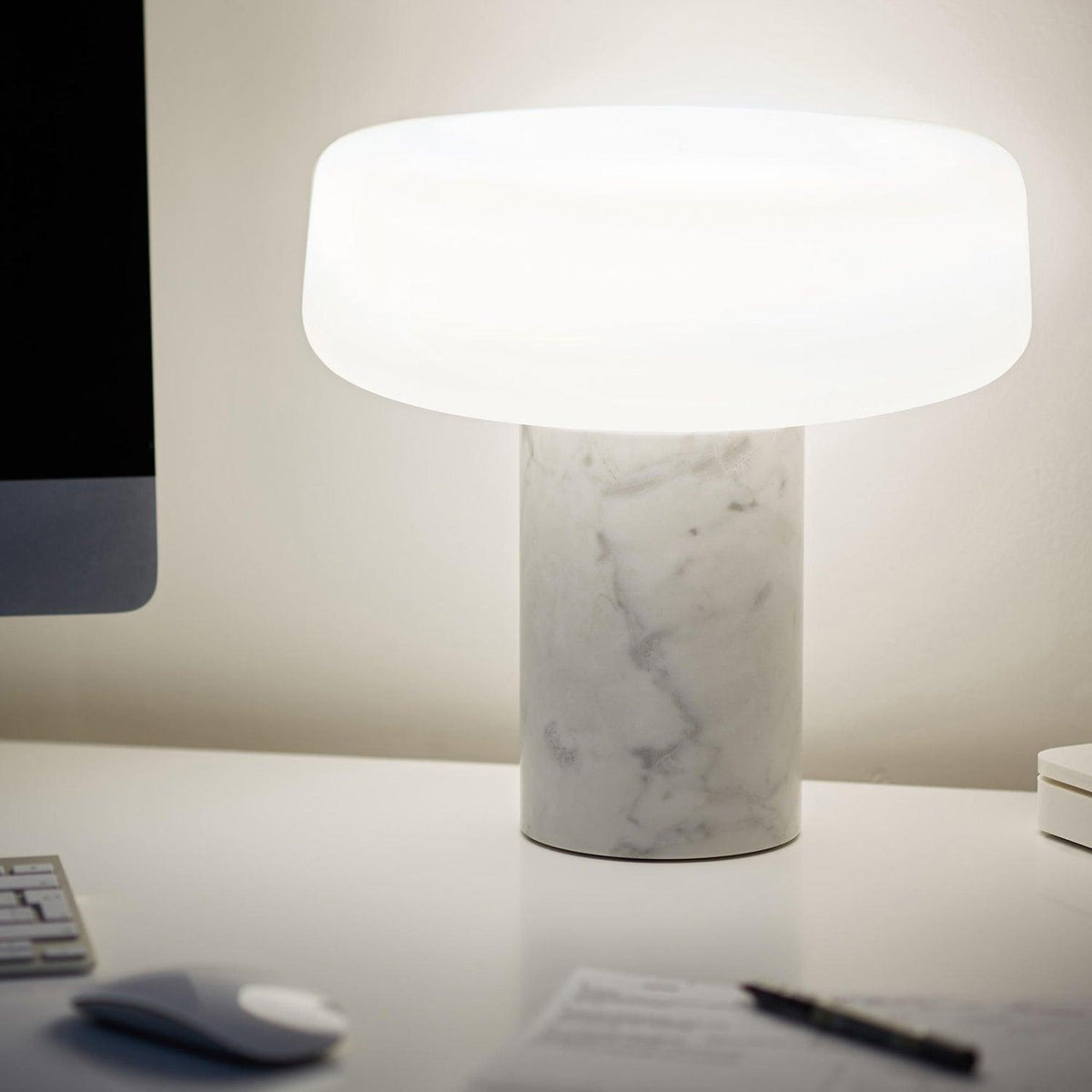 White marble Clérachic table lamp lit on a white office desk beside a computer monitor, a mouse and a pen.