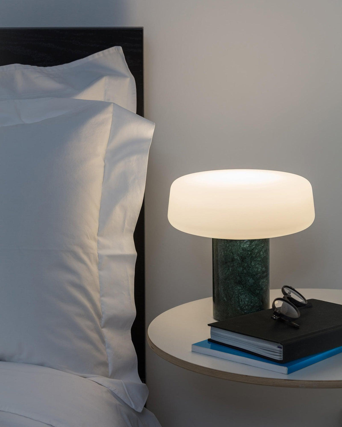 Green marble Clérachic table lamp glowing on a small round nightstand, framed by white bedding and pillows.