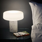 A book and reading glasses sit beside a white marble Clérachic table lamp lit on a glass bedside table.