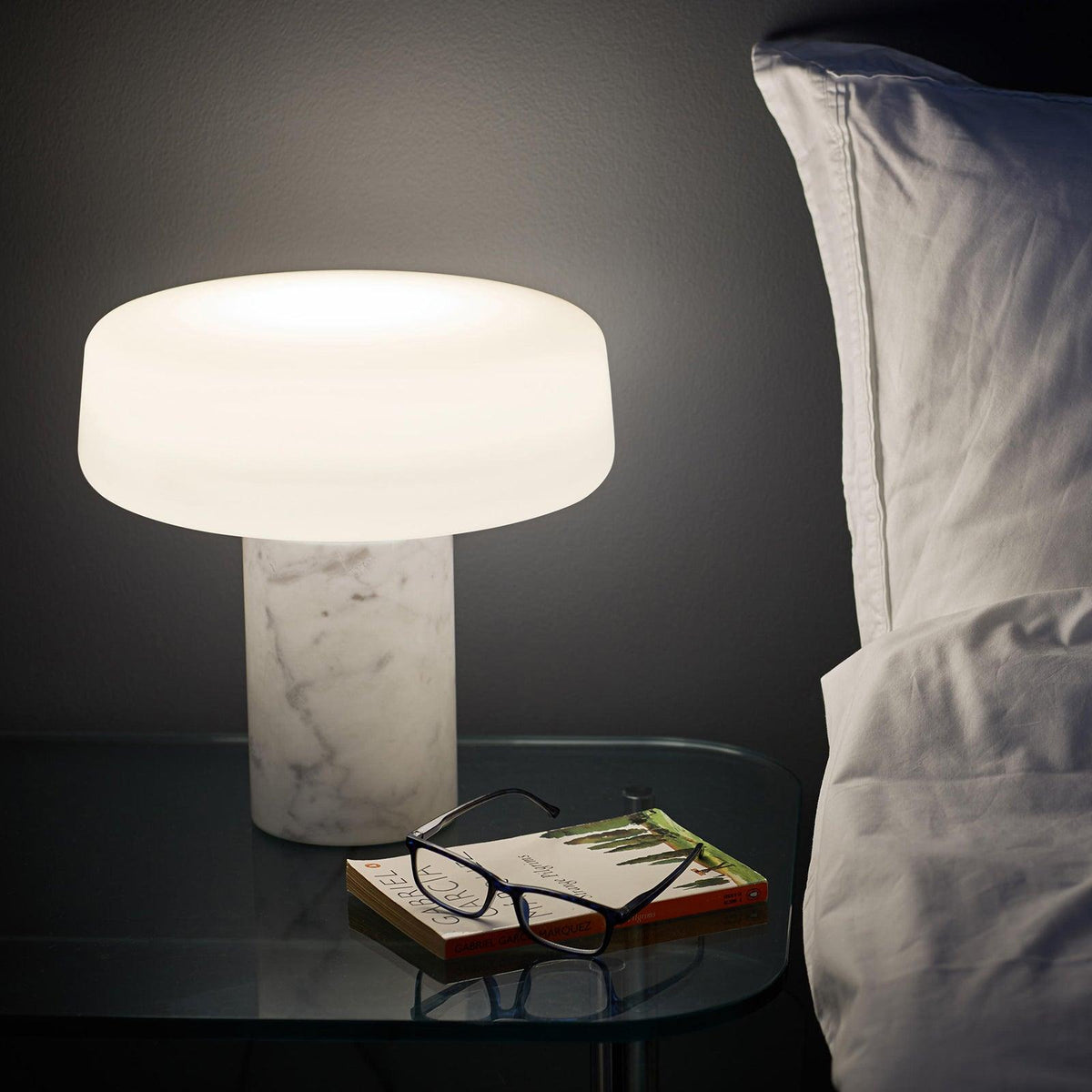 A book and reading glasses sit beside a white marble Clérachic table lamp lit on a glass bedside table.