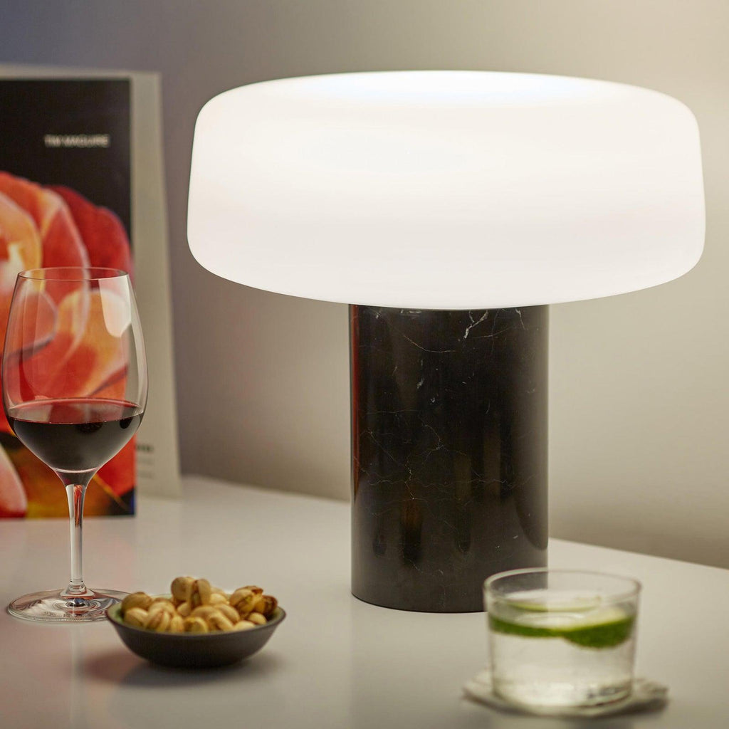 Black marble Clérachic table lamp lit on a white counter beside a glass of red wine, a bowl of nuts and a magazine.