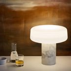 White marble Clérachic table lamp glowing on a wooden sideboard, a carafe and two glasses set against a warm rust-toned wall.