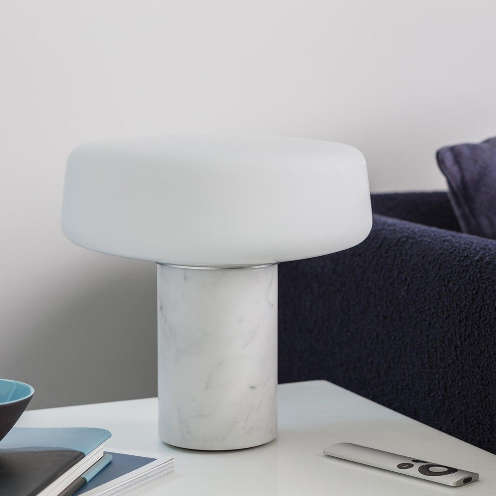 White marble Clérachic table lamp on a white desk with a small bowl, a notebook and a remote control close by.