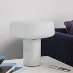 White marble Clérachic table lamp on a white desk with a small bowl, a notebook and a remote control close by.