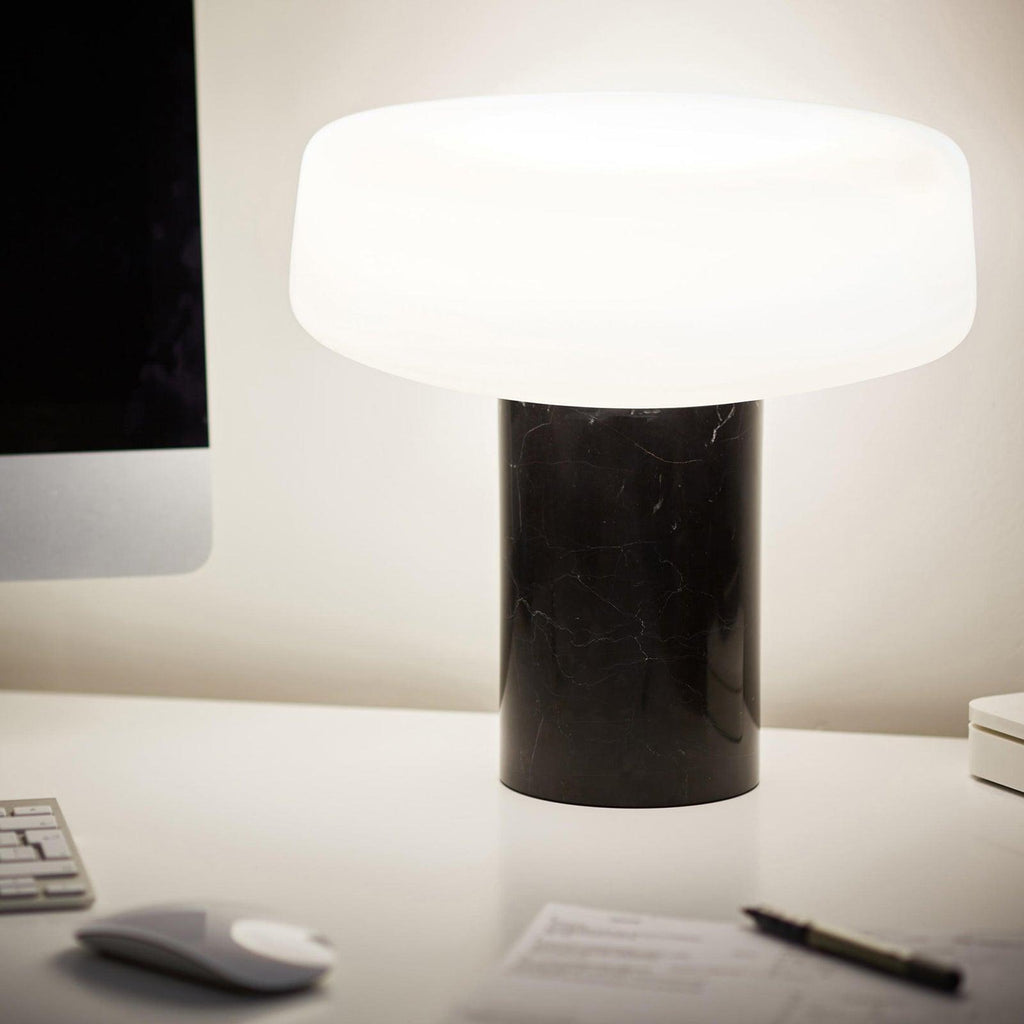 Black marble Clérachic table lamp lit on a white desk beside a computer monitor, a mouse and a pen, softly blurred behind.