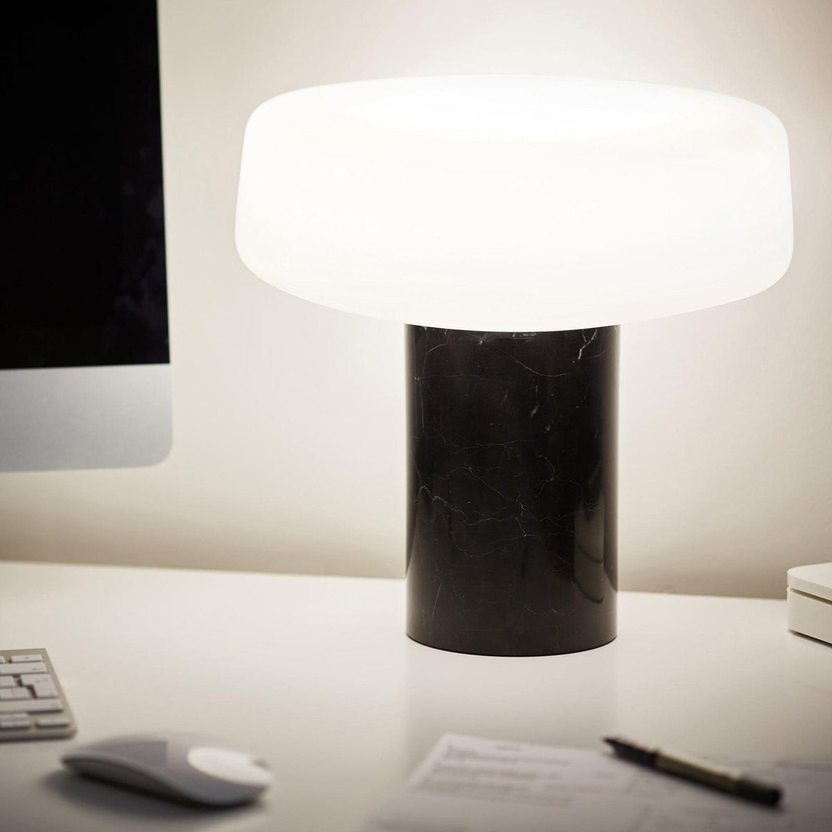 Black marble Clérachic table lamp lit on a white desk beside a computer monitor, a mouse and a pen, softly blurred behind.
