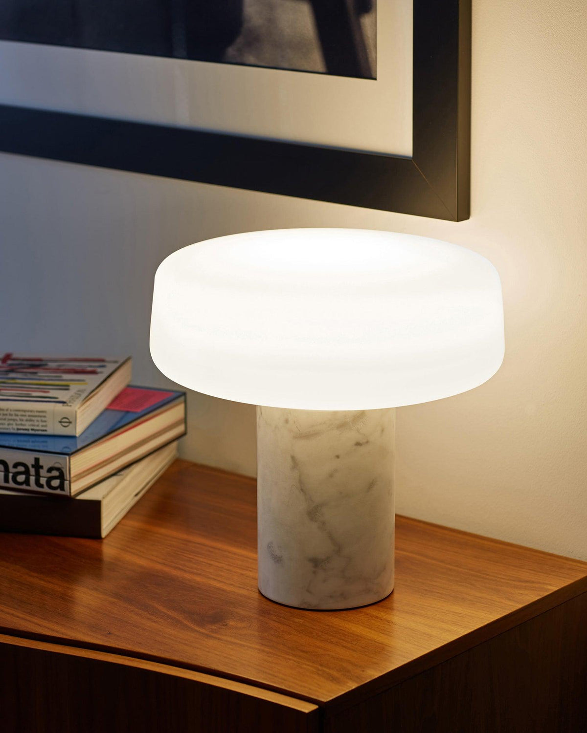 White marble Clérachic table lamp glowing on a wooden side table stacked with books, a framed picture on the wall behind.