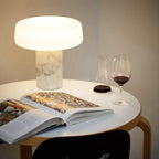 A glass of wine and a small carafe sit beside a white marble Clérachic table lamp lit on a round white table.