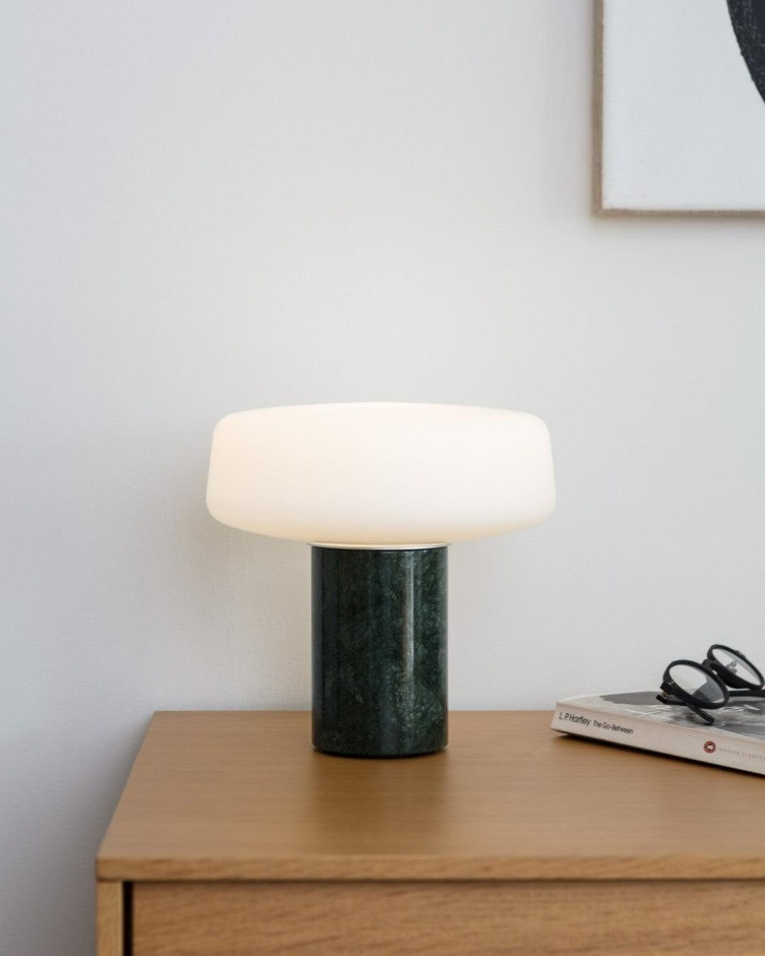 Reading glasses rest on a stack of books beside a green marble Clérachic table lamp lit on a wooden nightstand.