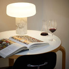 Seen from above, a white marble Clérachic table lamp lit on a round table sits beside an open magazine and wine.
