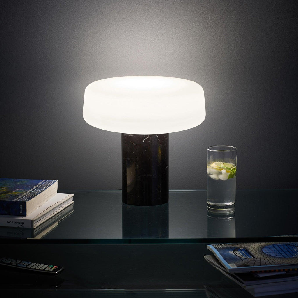 Black marble Clérachic table lamp glowing beside a glass of sparkling water and stacked books on a dark glass shelf.