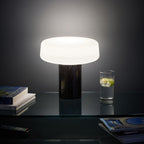Black marble Clérachic table lamp glowing beside a glass of sparkling water and stacked books on a dark glass shelf.