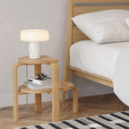 Books and glasses line the shelf below a two-tier wooden stool holding a lit white marble Clérachic table lamp beside a bed.