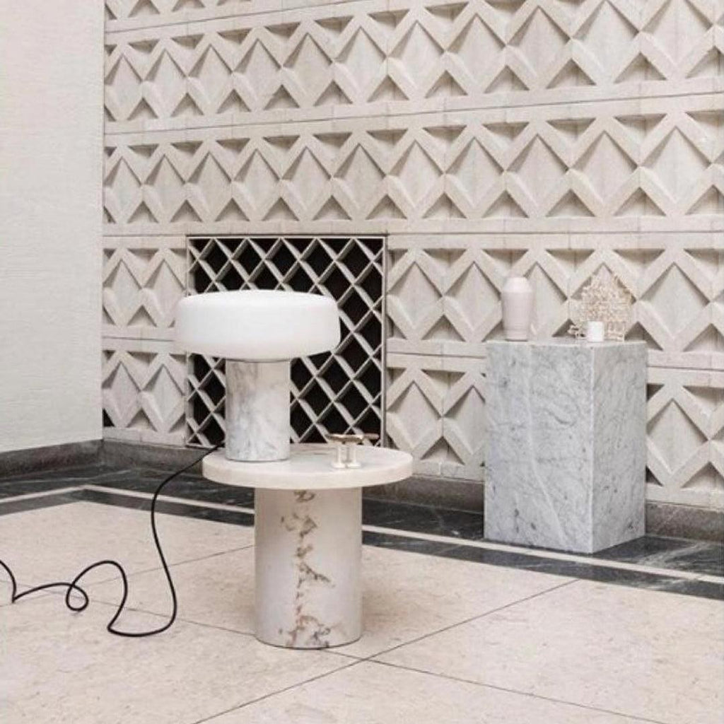 A marble plinth stands alongside a white marble Clérachic table lamp lit on a round pedestal table against tiled walls.