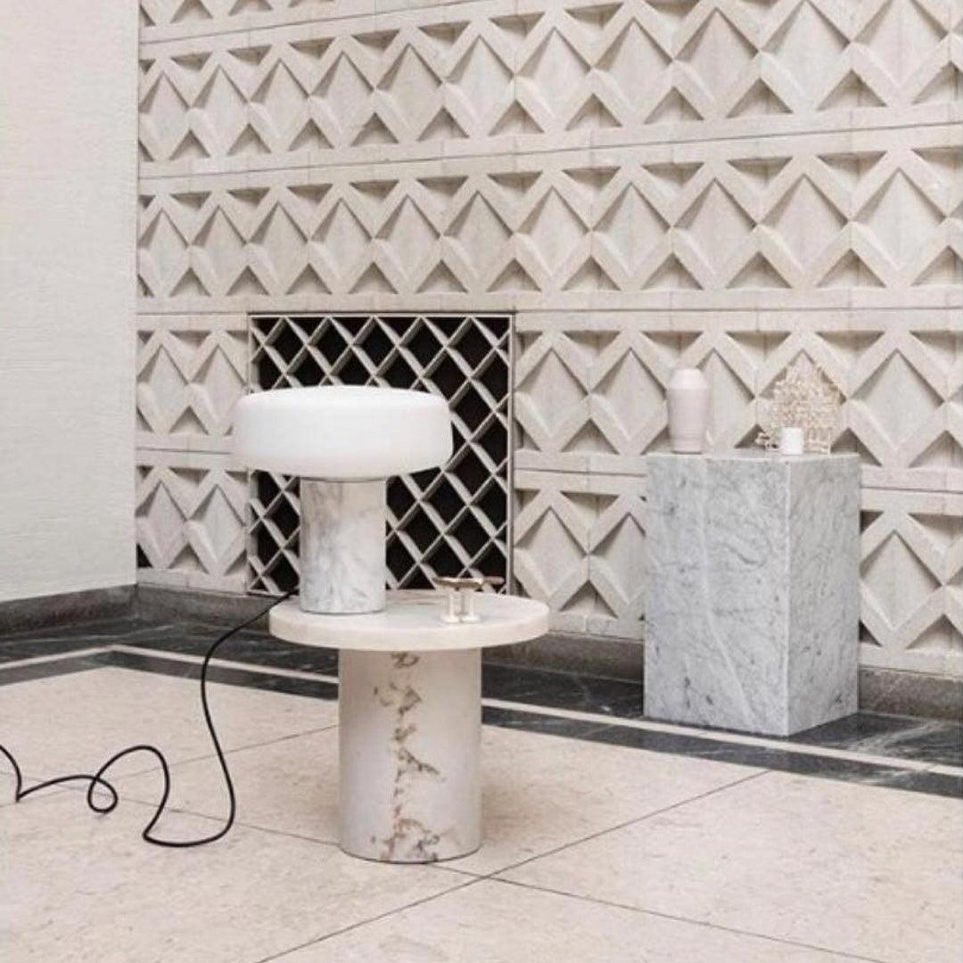 A marble plinth stands alongside a white marble Clérachic table lamp lit on a round pedestal table against tiled walls.