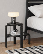 A dark bed frame stands beside a two-tier stool holding books, glasses and a lit black marble Clérachic table lamp.