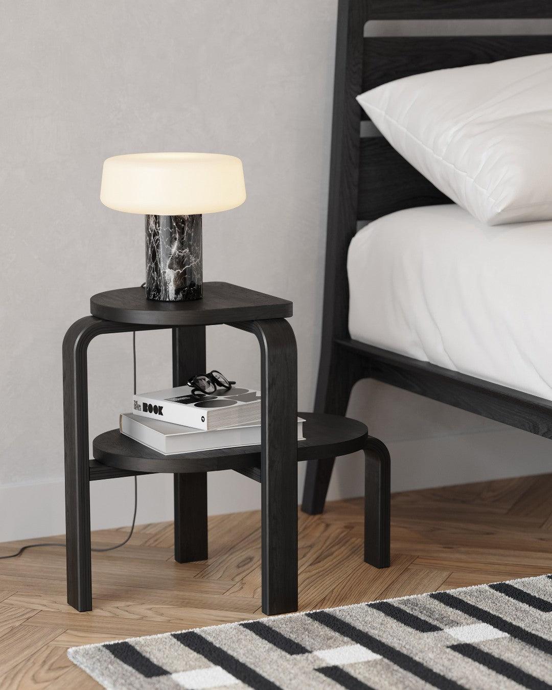 A dark bed frame stands beside a two-tier stool holding books, glasses and a lit black marble Clérachic table lamp.