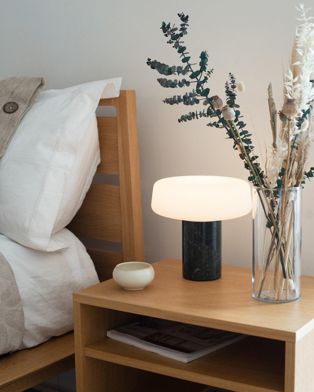 Dark marble Clérachic table lamp lit on a wooden nightstand next to eucalyptus stems in a glass vase and a small bowl.