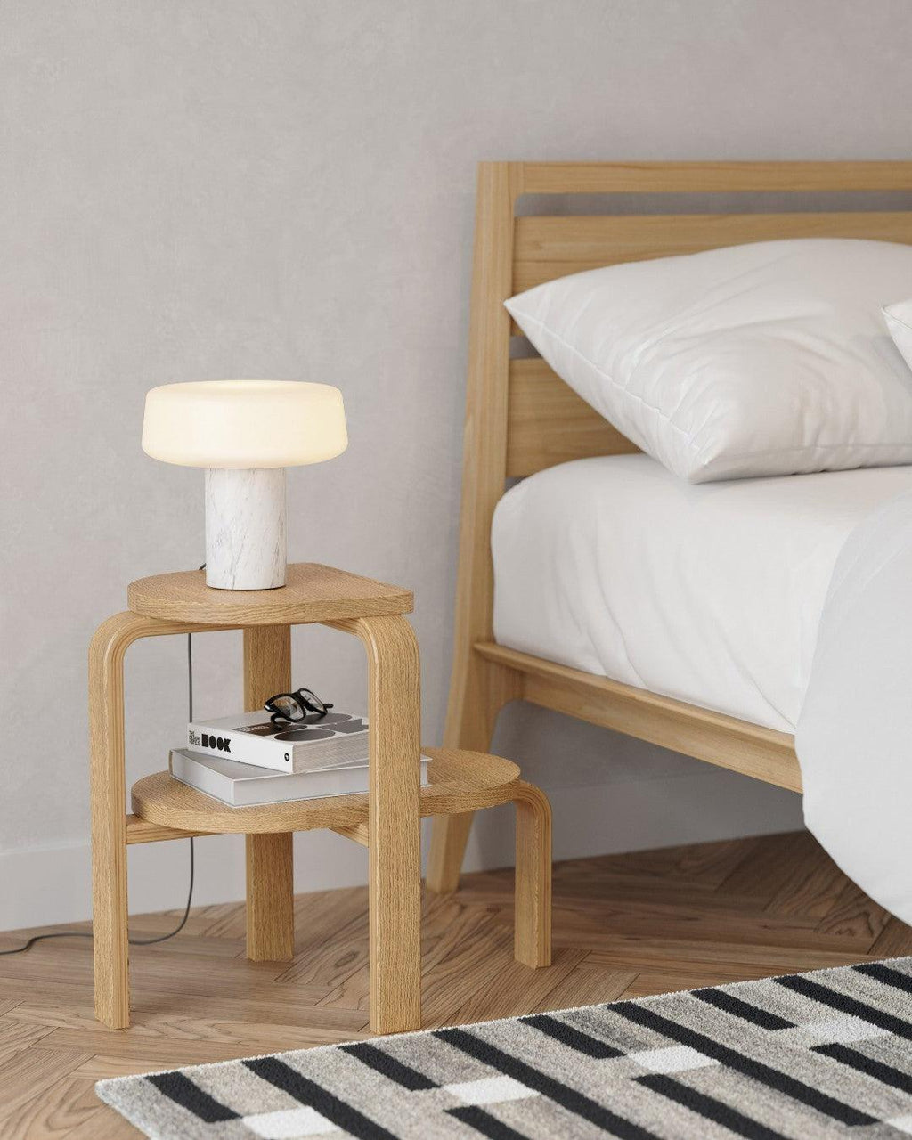 White pillows dress a bed beside a lit white marble Clérachic table lamp atop a wooden stool, books stacked below.