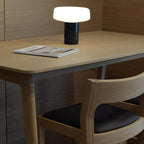 An open magazine lies on the desk beside a wooden chair and a black marble Clérachic table lamp lit nearby.