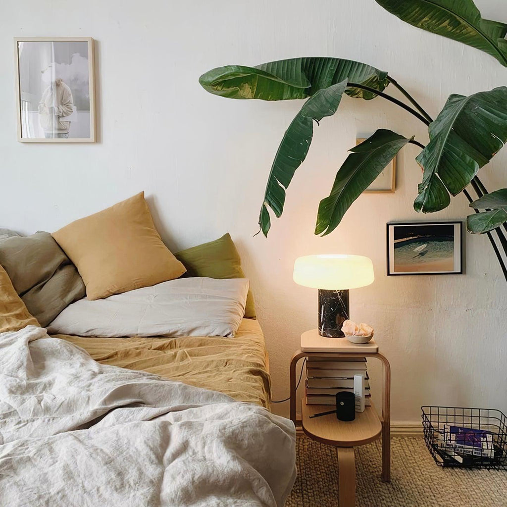 Dark marble Clérachic table lamp lit on a bedside table with mustard and cream pillows, a leafy print framed on the wall.