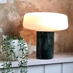 A trailing houseplant sits beside a green marble Clérachic table lamp lit on a stone counter against a weathered rust wall.