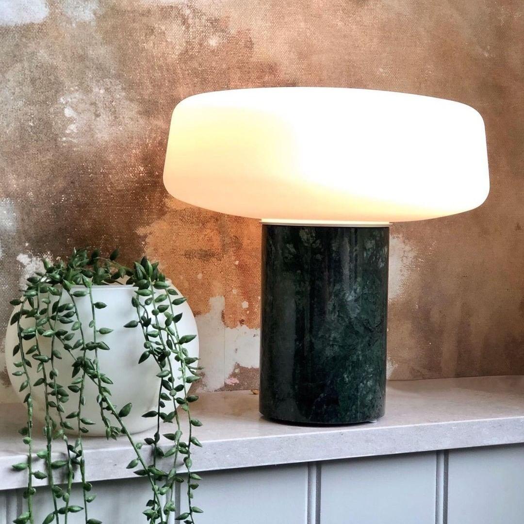 A trailing houseplant sits beside a green marble Clérachic table lamp lit on a stone counter against a weathered rust wall.