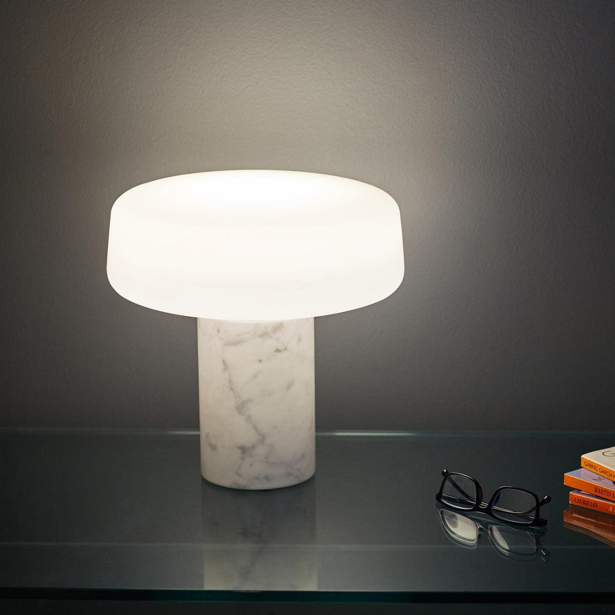 White marble Clérachic table lamp casting a warm glow on a dark glass shelf stacked with books and reading glasses.