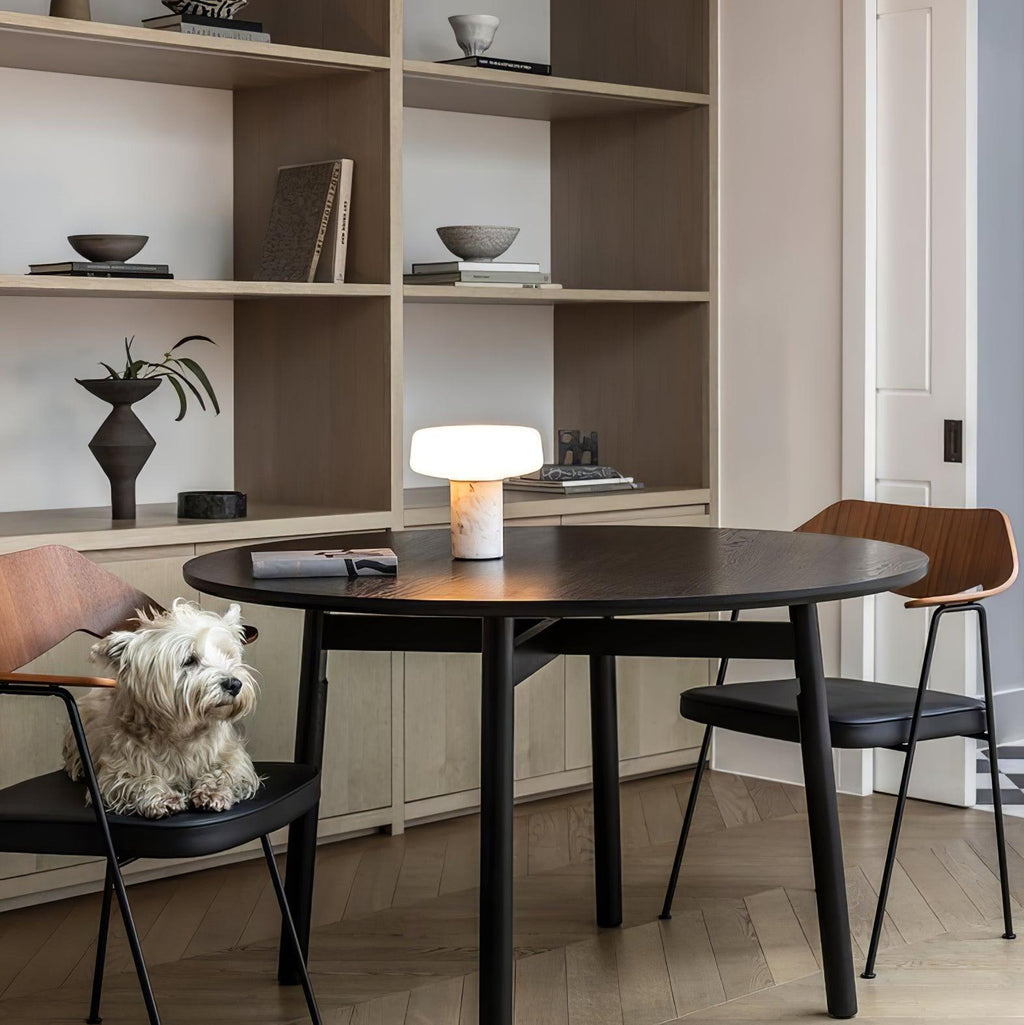 A small dog perches on a chair behind a white marble Clérachic table lamp lit on a round dining table.