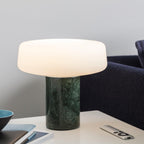 A small blue bowl and remote lie beside a green marble Clérachic table lamp lit near a navy sofa.
