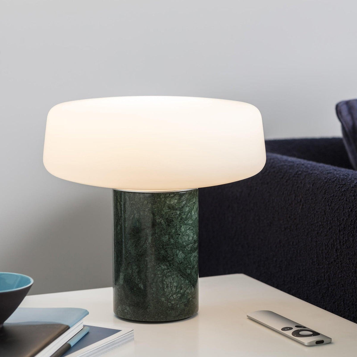 A small blue bowl and remote lie beside a green marble Clérachic table lamp lit near a navy sofa.