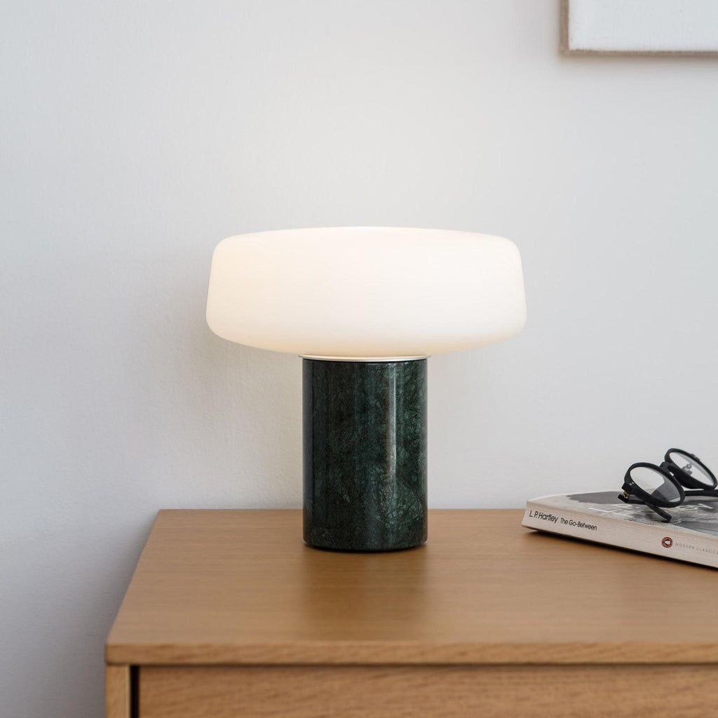A stack of books sits beside reading glasses and a green marble Clérachic table lamp lit on a wooden nightstand.