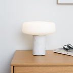 White marble Clérachic table lamp lit on a wooden bedside dresser, with reading glasses and a magazine nearby.
