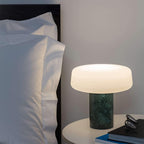 Green marble Clérachic table lamp lit on a round nightstand beside a bed with white pillows and a stack of books.