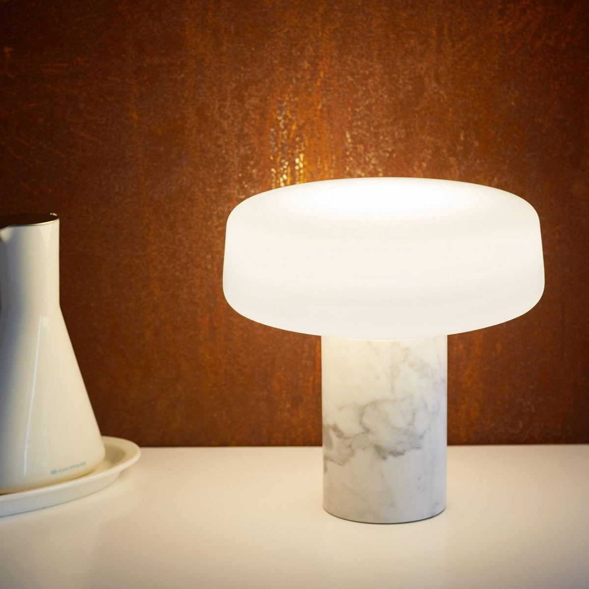 White marble Clérachic table lamp glowing on a white counter in front of a weathered copper-toned wall, a ceramic carafe alongside.