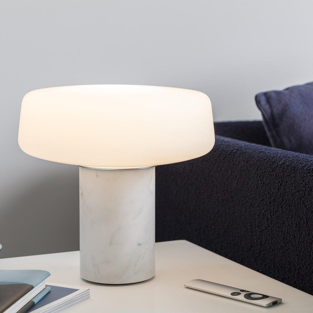 White marble Clérachic table lamp lit on a white side table in front of a navy sofa, with books and a remote control.