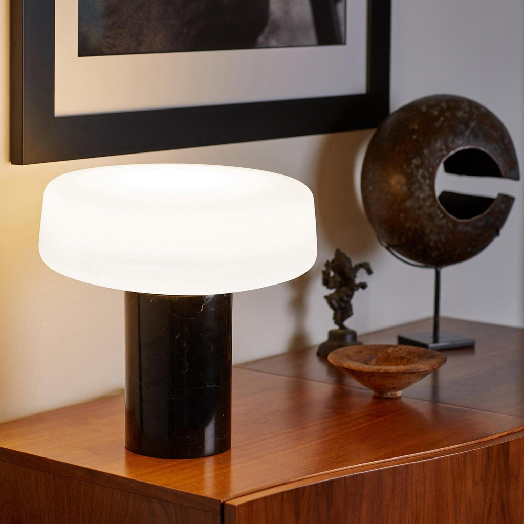 Black marble Clérachic table lamp lit on a wooden sideboard beside a framed print and an open-circle metal sculpture.