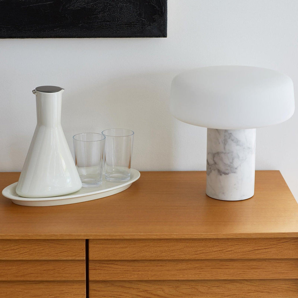 White marble Clérachic table lamp on a wooden dresser next to a ceramic carafe and two drinking glasses on a tray.