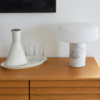 White marble Clérachic table lamp on a wooden dresser next to a ceramic carafe and two drinking glasses on a tray.