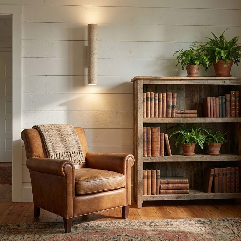 A brown leather armchair and bookshelf sit below this Clérachic wall light, lit, in a rustic living room.