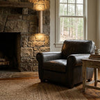 A stone fireplace and black leather armchair sit near this Clérachic wall light, lit, in a living room.
