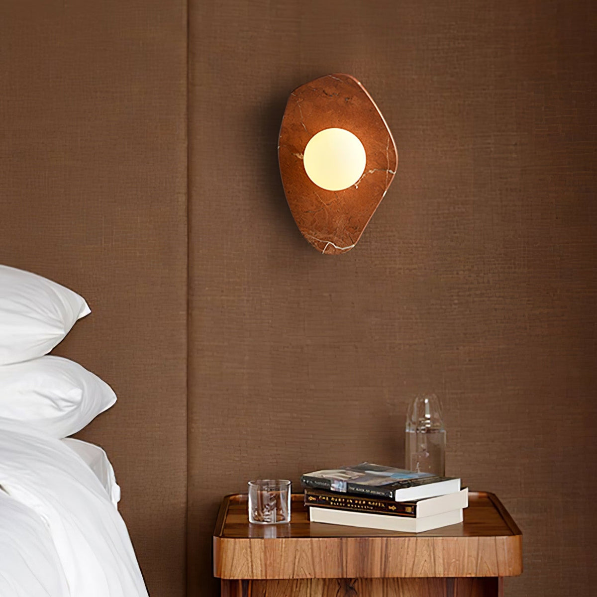 This Clérachic wall light glows above a rust headboard, beside a walnut nightstand stacked with books and a carafe.