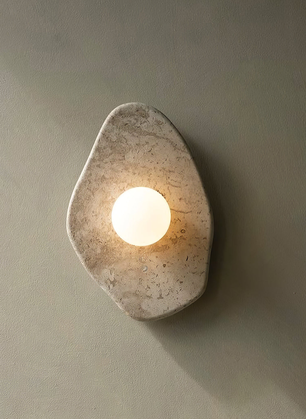 The beige stone wall light from Clérachic glowing on a sage-grey wall, casting a soft shadow beneath it.
