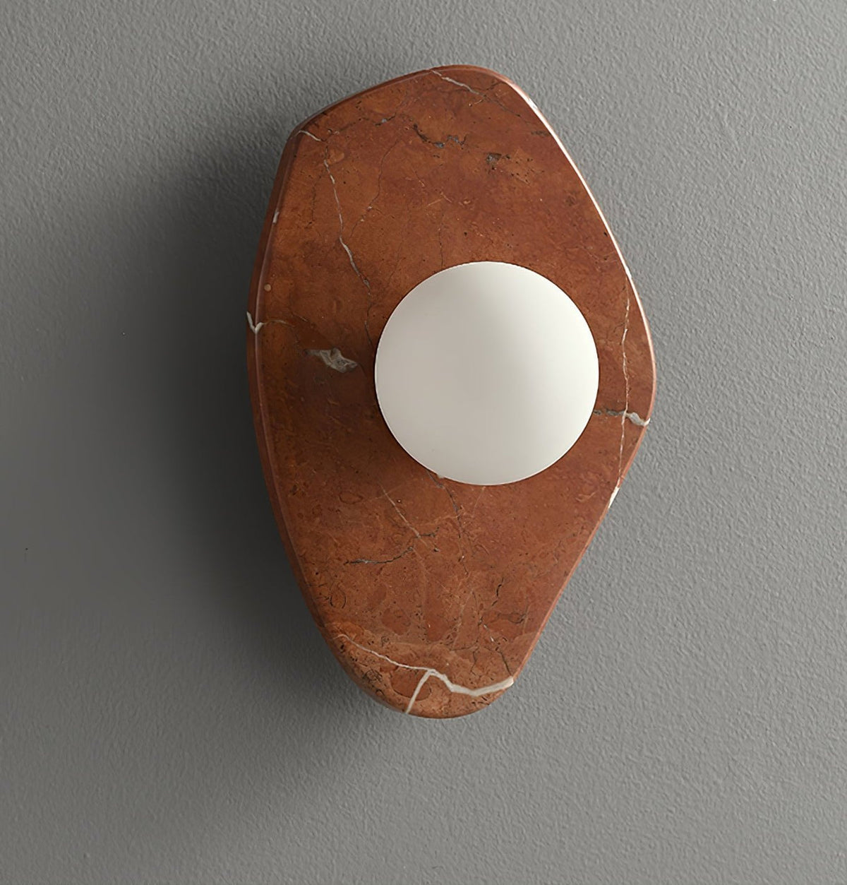 Wide view of the coral-red marble wall light from Clérachic, lit, on a grey studio wall with the orb centred low.