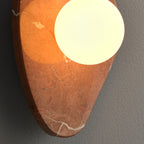 Angled close-up of the coral-red marble wall light from Clérachic, the white glass orb glowing against warm veining.